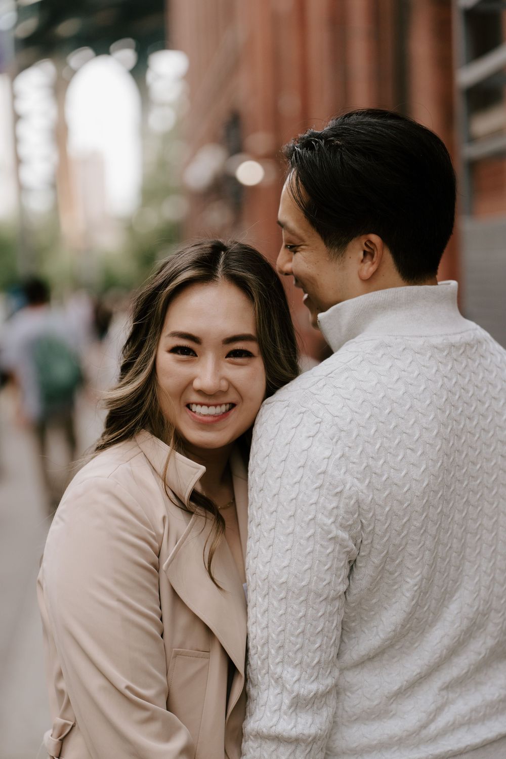 A smiling Asian woman in a beige coat leans close to a man in a white sweater on a city street.