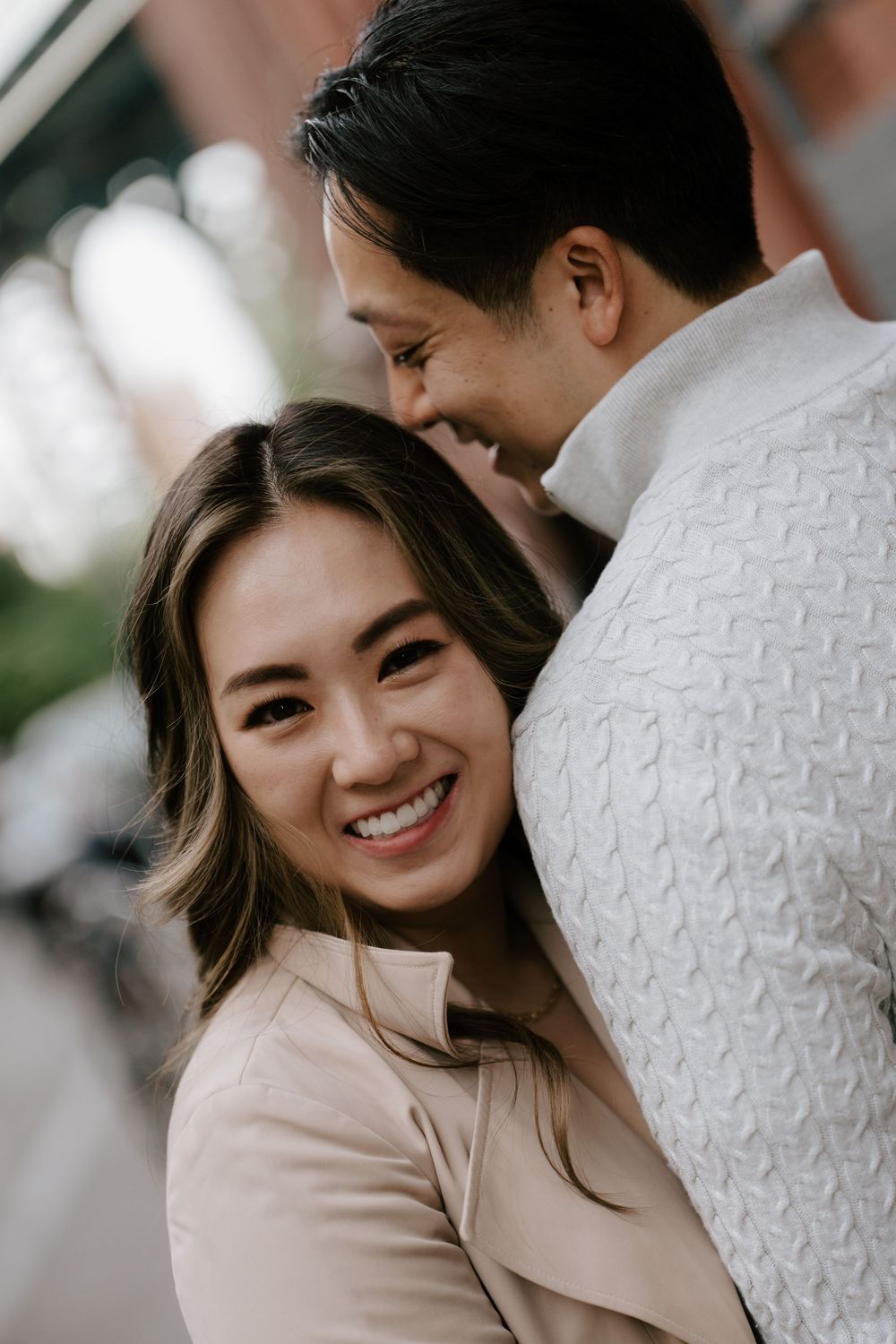 A smiling Asian woman in a beige jacket embraces a man in a white sweater during an outdoor couples photo session.