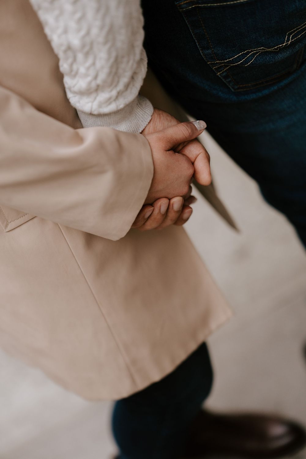 Two people holding hands tenderly, one wearing a beige blazer and white sweater, the other in dark blue jeans.