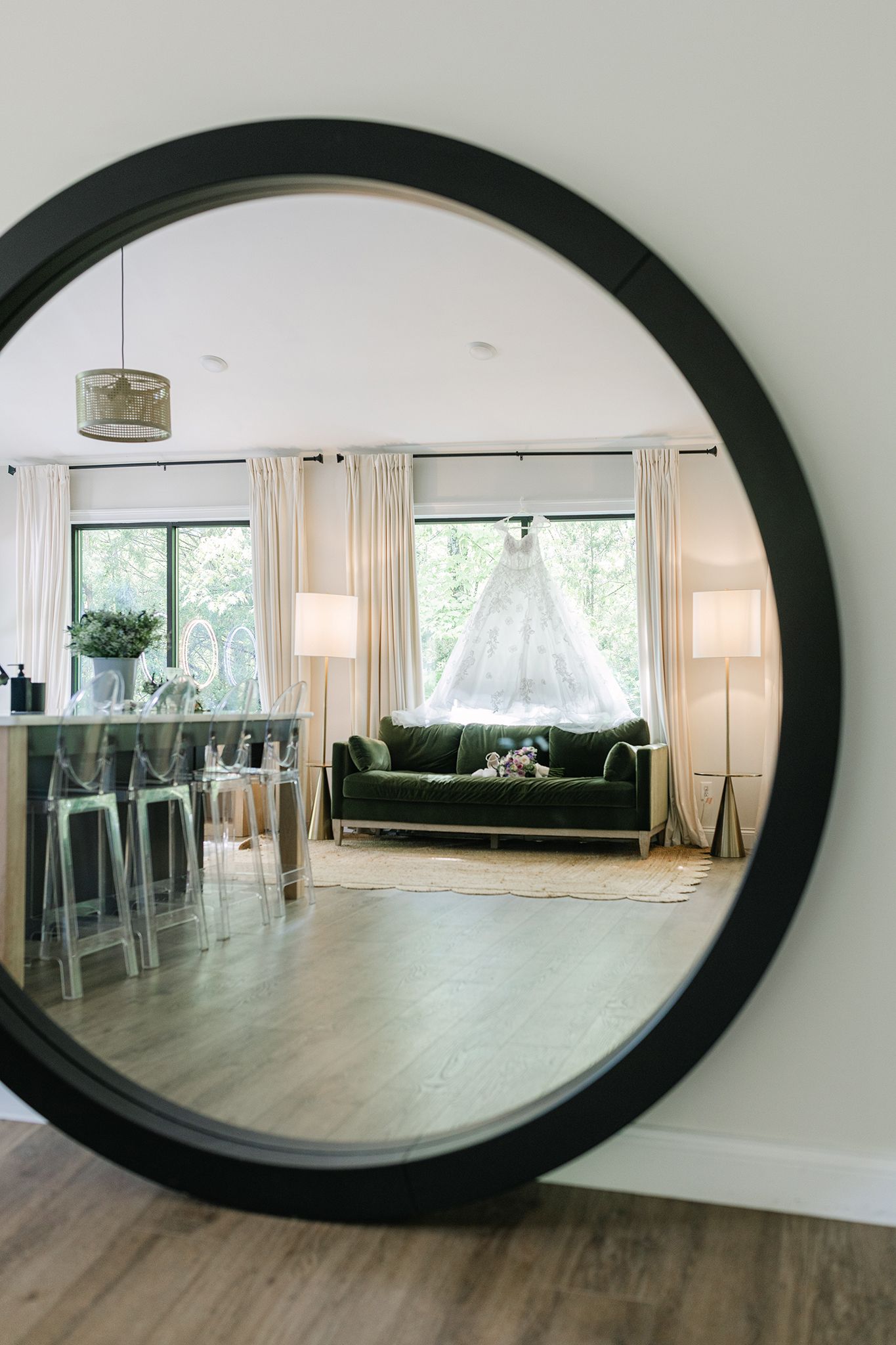 Round black mirror reflecting a bridal suite with a wedding dress hanging by a window above a green velvet sofa.