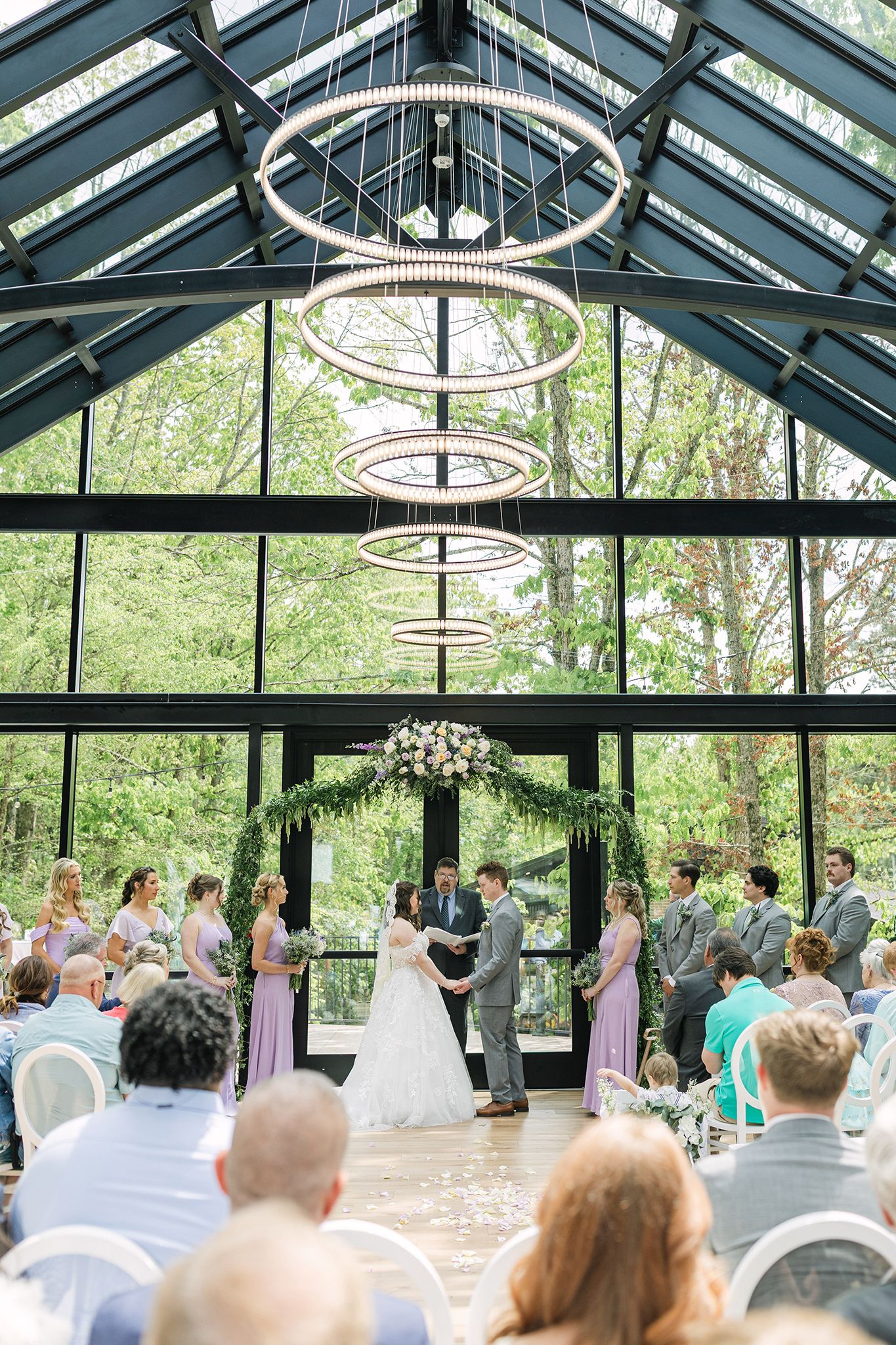 Elegant greenhouse wedding ceremony with circular chandelier, floral arch, and lush greenery backdrop.