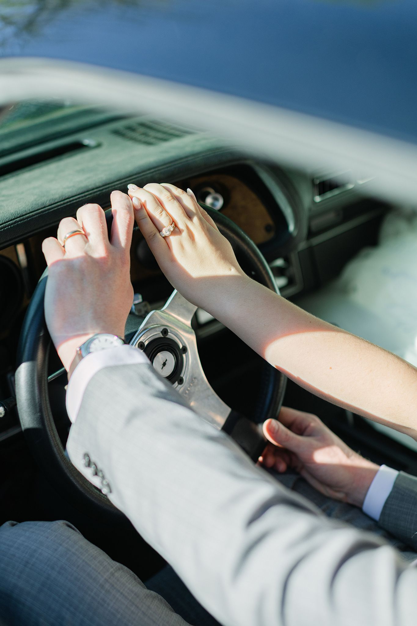 Newlywed couple holding hands on vintage car steering wheel, showing wedding rings, romantic moment inside classic vehicle.
