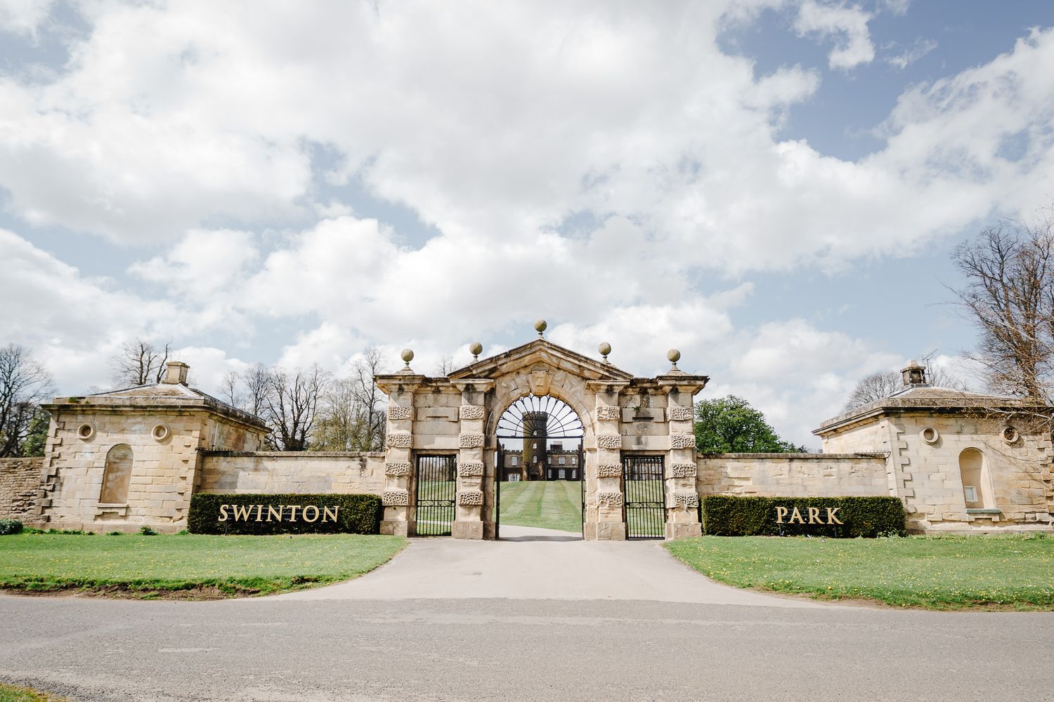 Stone gatehouse entrance to Swinton Park estate with ornate iron gates, manicured hedges, and cloudy sky backdrop.