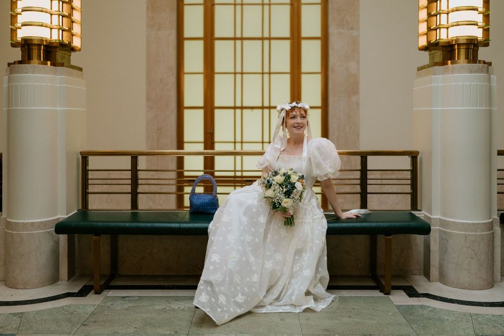 Bride in white lace gown with puff sleeves holds bouquet while seated on green bench in elegant Art Deco lobby.