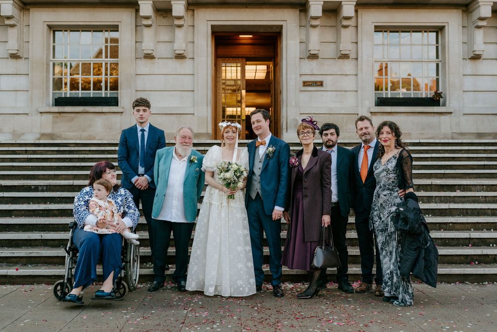 Wedding party group photo on steps outside a building, bride in lace gown with bouquet, groom in teal suit.