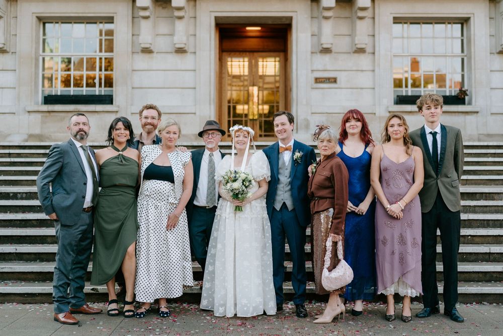 Wedding party poses on stone steps outside a grand building, bride in white gown with bouquet surrounded by family.