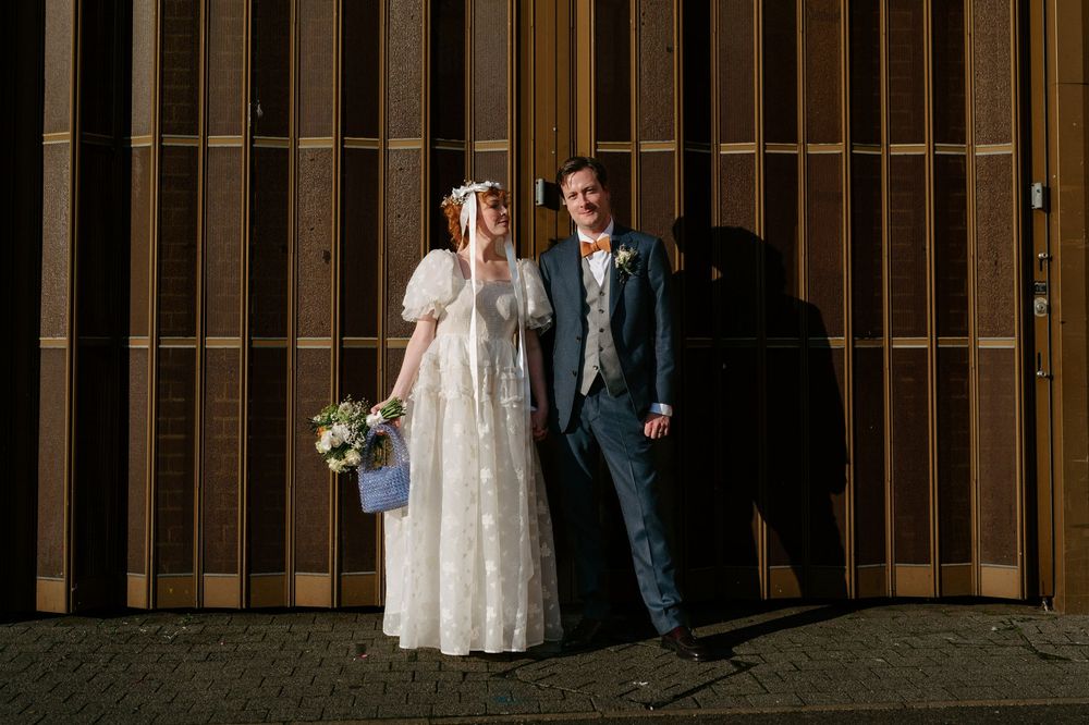 Bride and groom pose together laughing against wooden wall in natural sunlight.