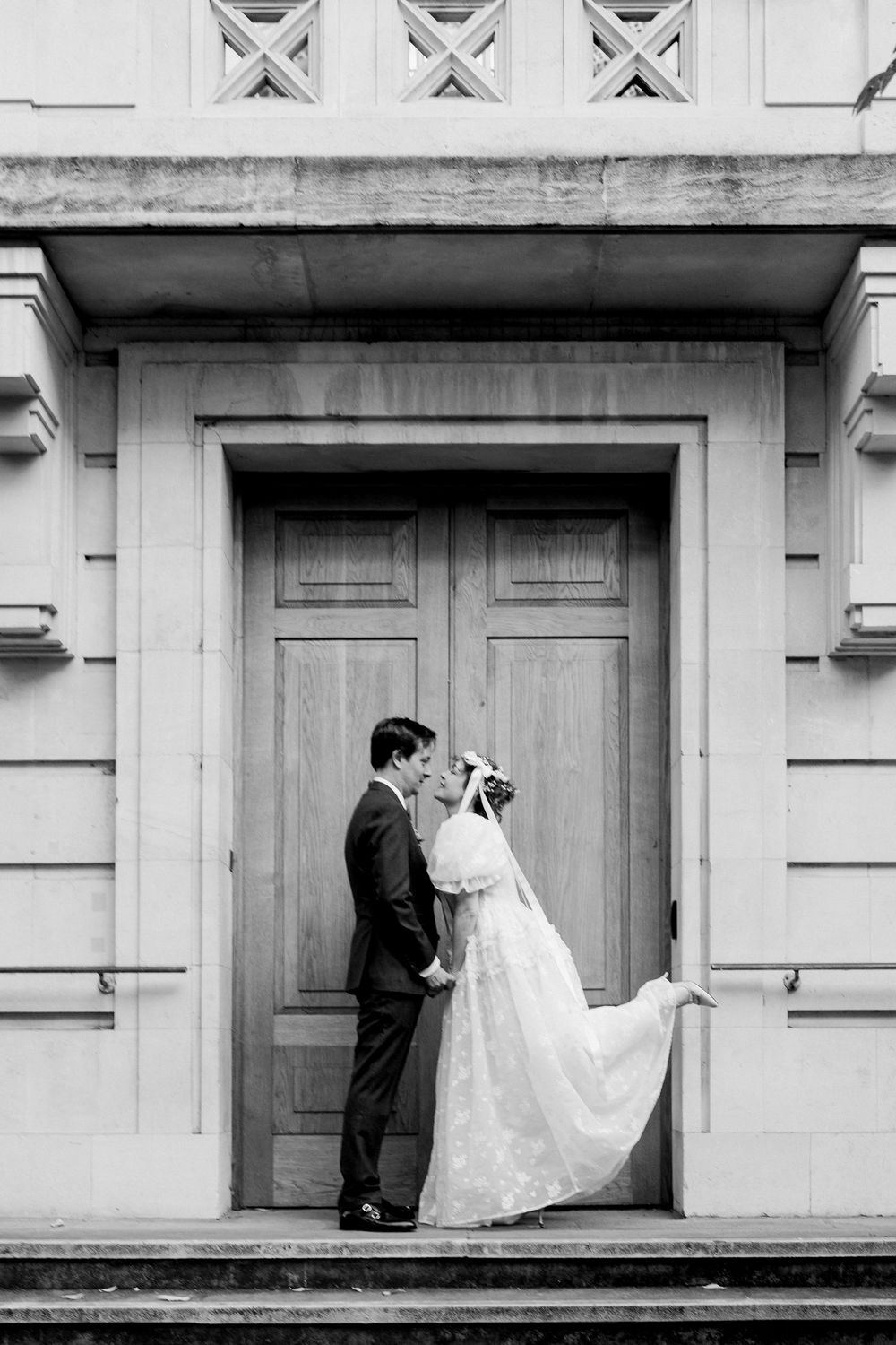 Black and white photo of bride and groom sharing a kiss in front of elegant wooden doors on stone steps.