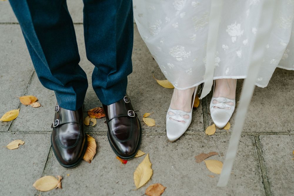 Close-up of bride and groom shoes on autumn cobblestones, dark monk-strap oxfords and white bridal heels.