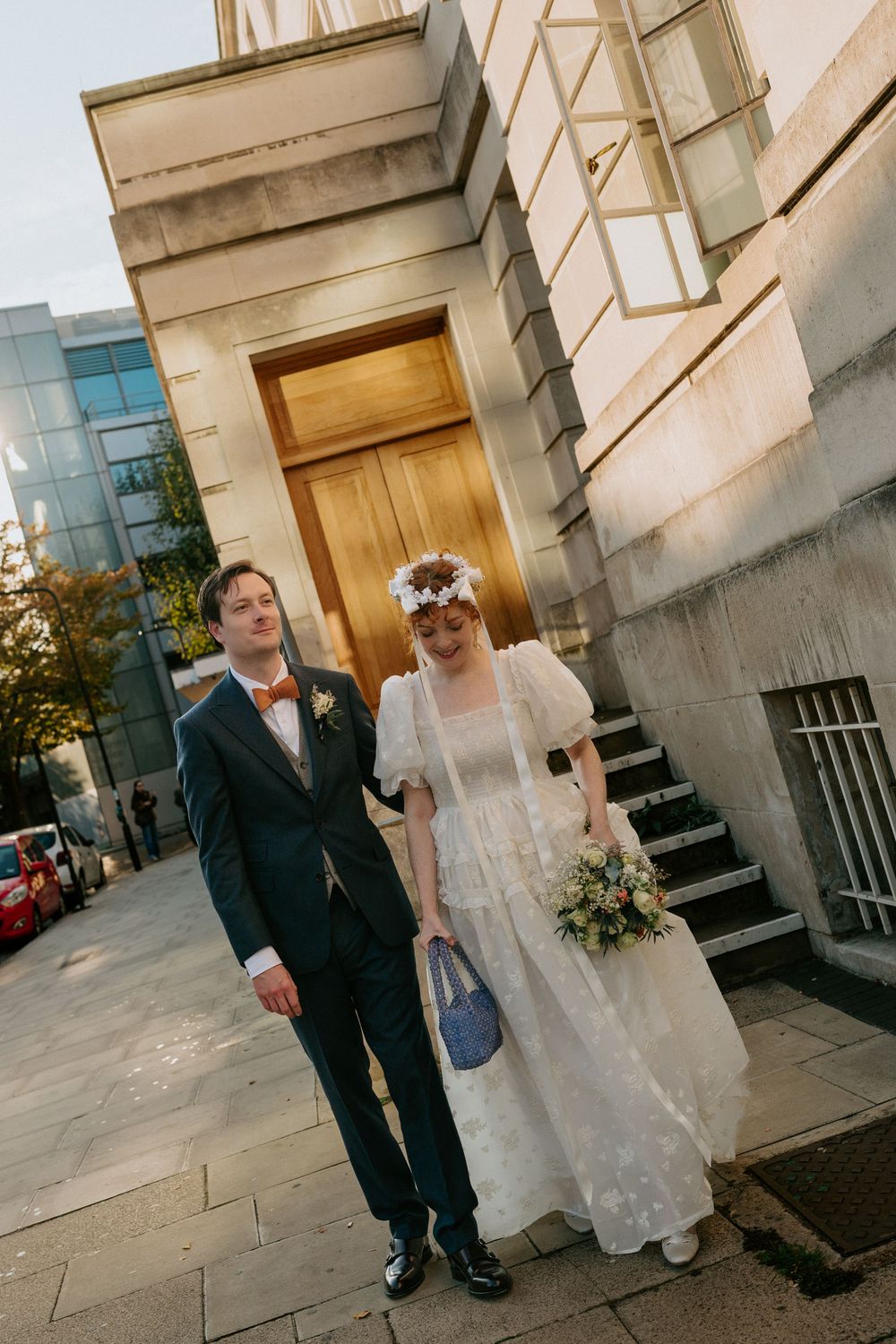 Bride and groom walking outside historic stone building at golden hour, bride in lace dress with floral crown.