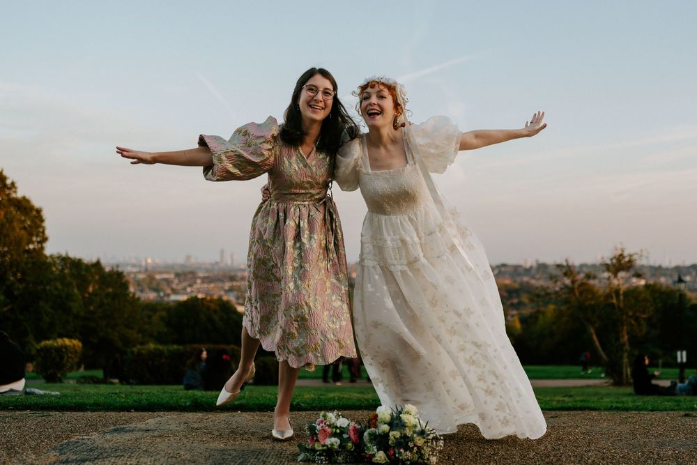 Bride and bridesmaid joyfully pose with arms outstretched at sunset with city skyline view.