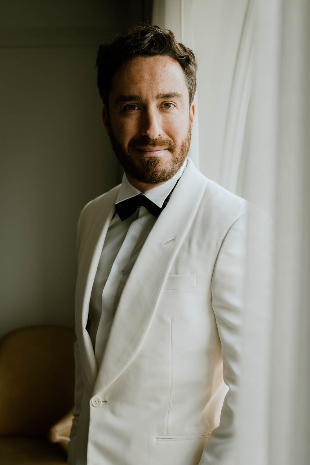 A man in a white tuxedo jacket and black bow tie poses confidently near a window with soft natural light.