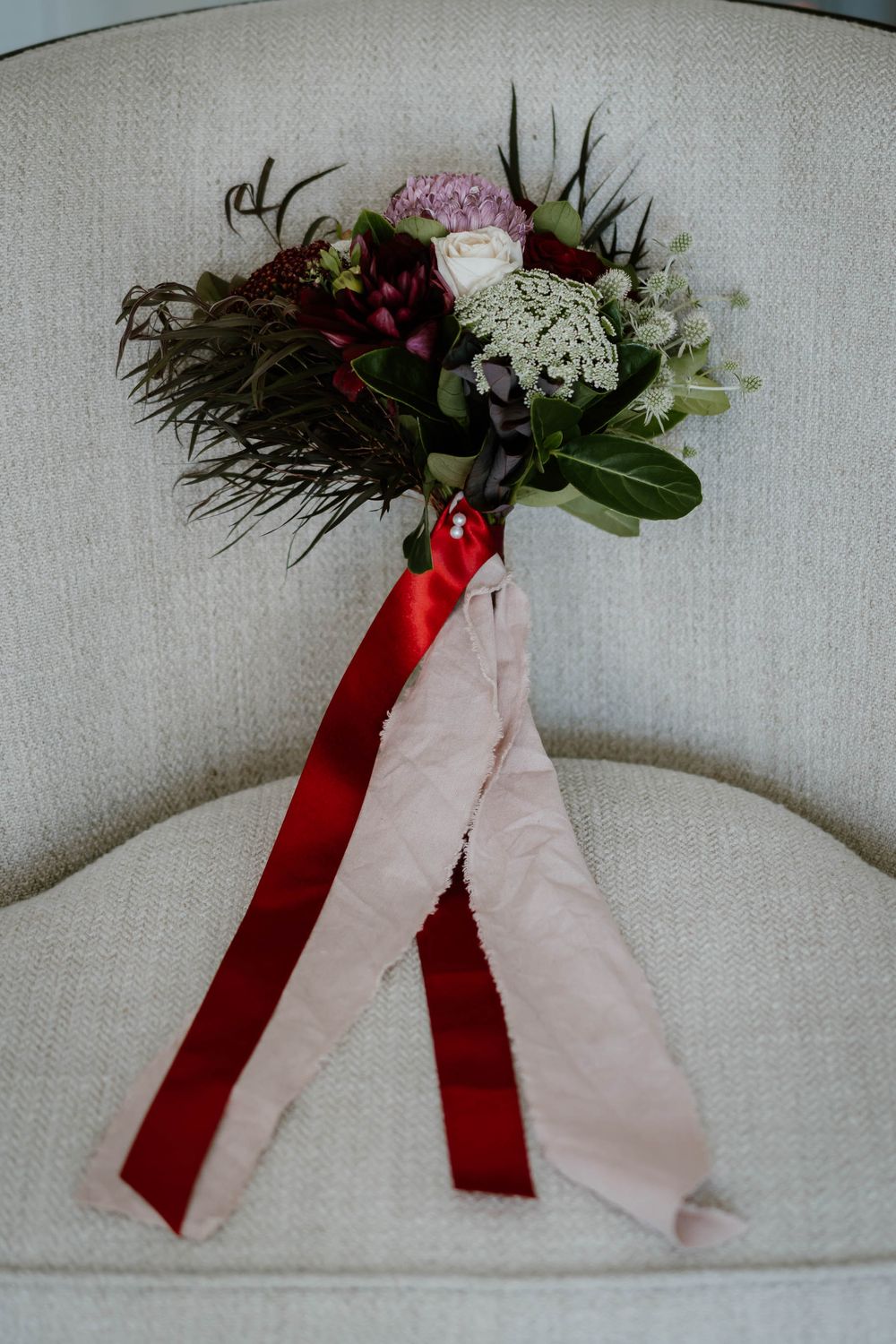 Rustic wedding bouquet with burgundy and blush flowers, greenery, and red and pink satin ribbons resting on a white chair.