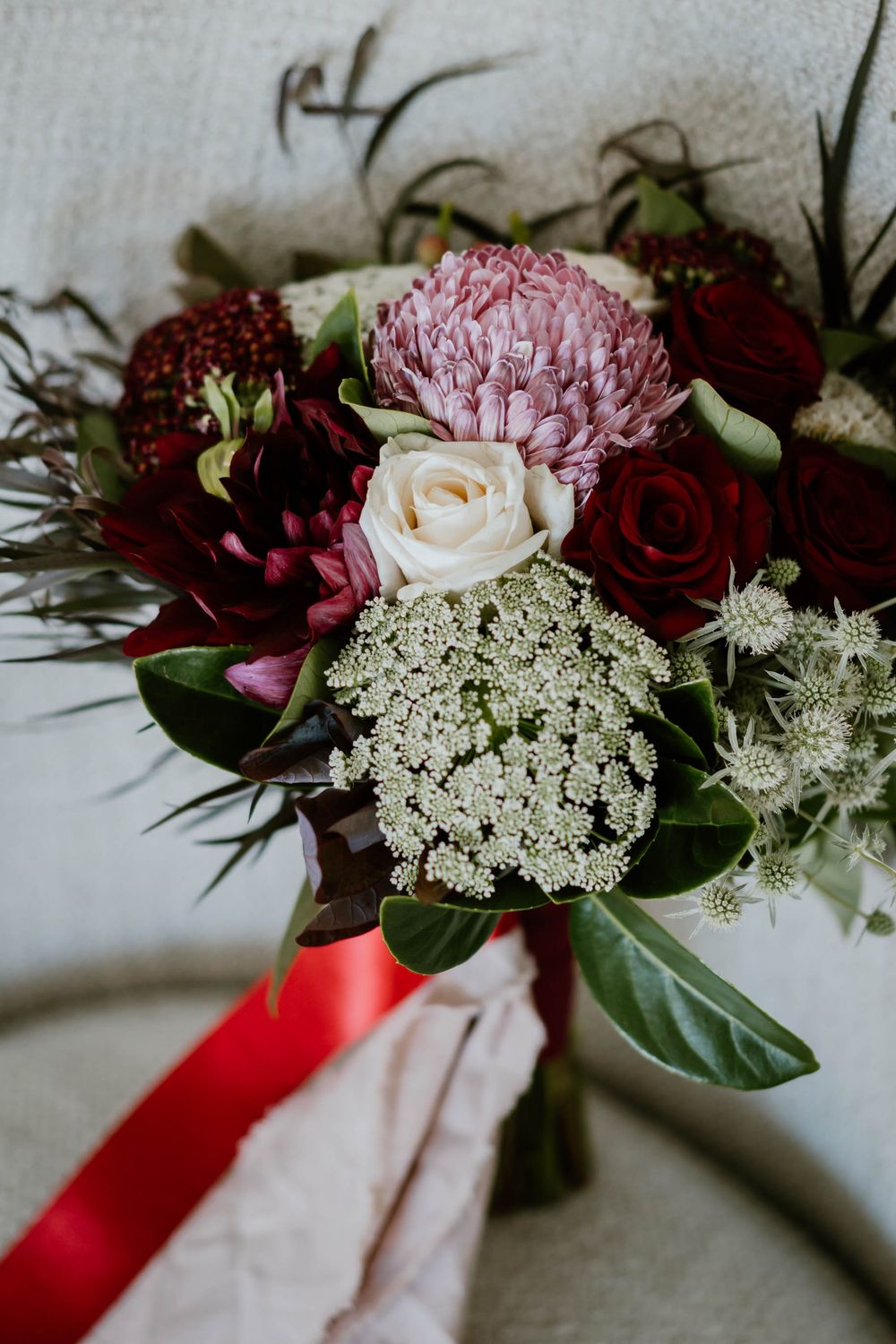 Elegant bridal bouquet featuring burgundy roses, pink protea, white blooms, and greenery tied with a red ribbon.