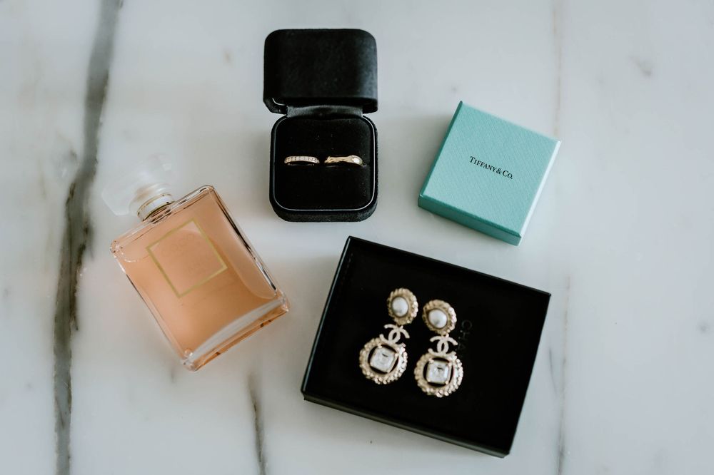 Chanel perfume bottle, diamond ring in black box, Tiffany box, and pearl Chanel earrings on marble surface.