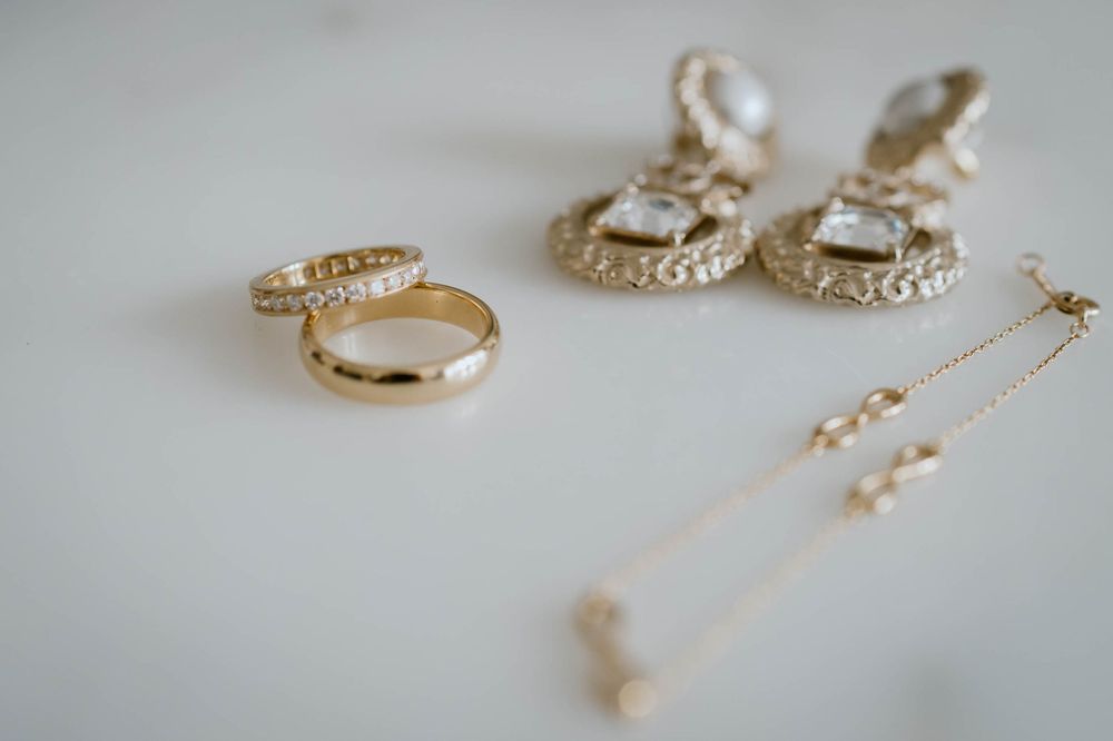 Gold wedding rings, ornate crystal drop earrings, and a delicate pearl chain necklace arranged on a white surface.