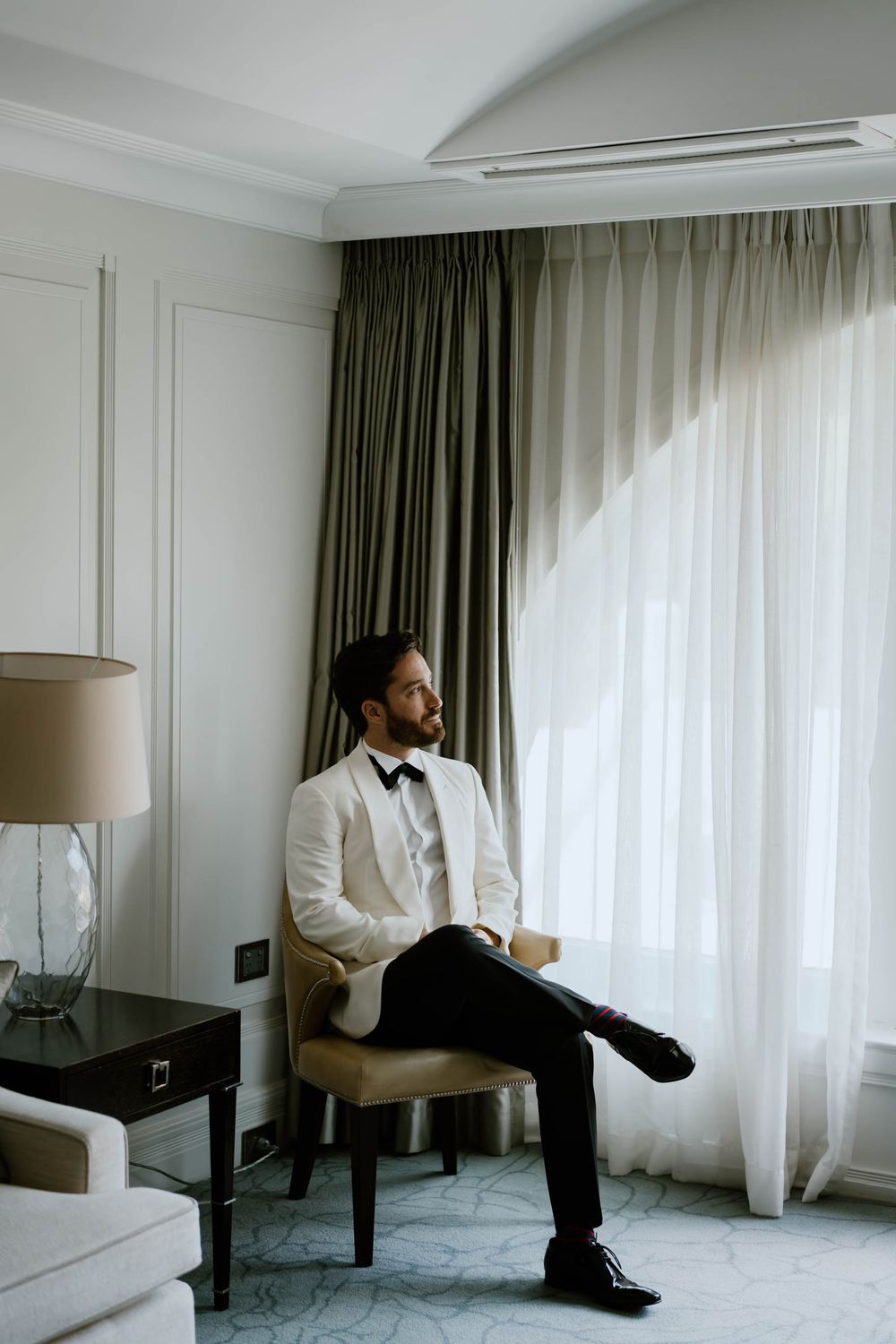 A well-dressed groom in a white blazer and bow tie sits cross-legged on a chair in an elegant hotel room.