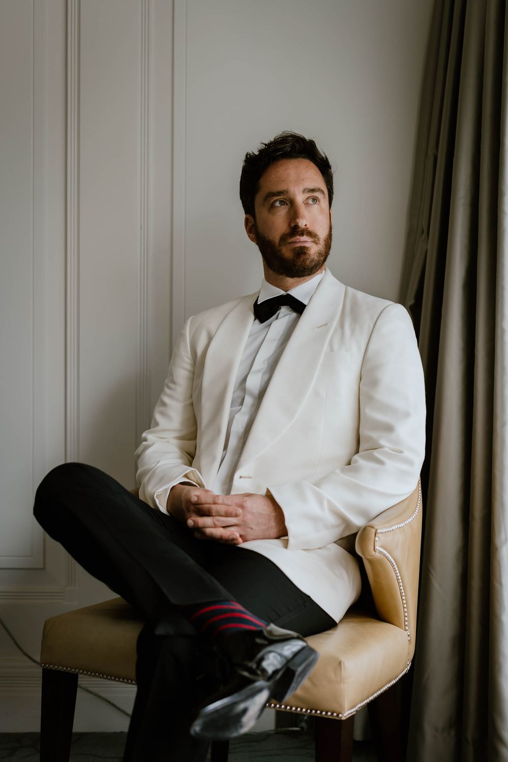 A dapper groom in a white tuxedo jacket and bow tie sits cross-legged in a chair, gazing thoughtfully upward.