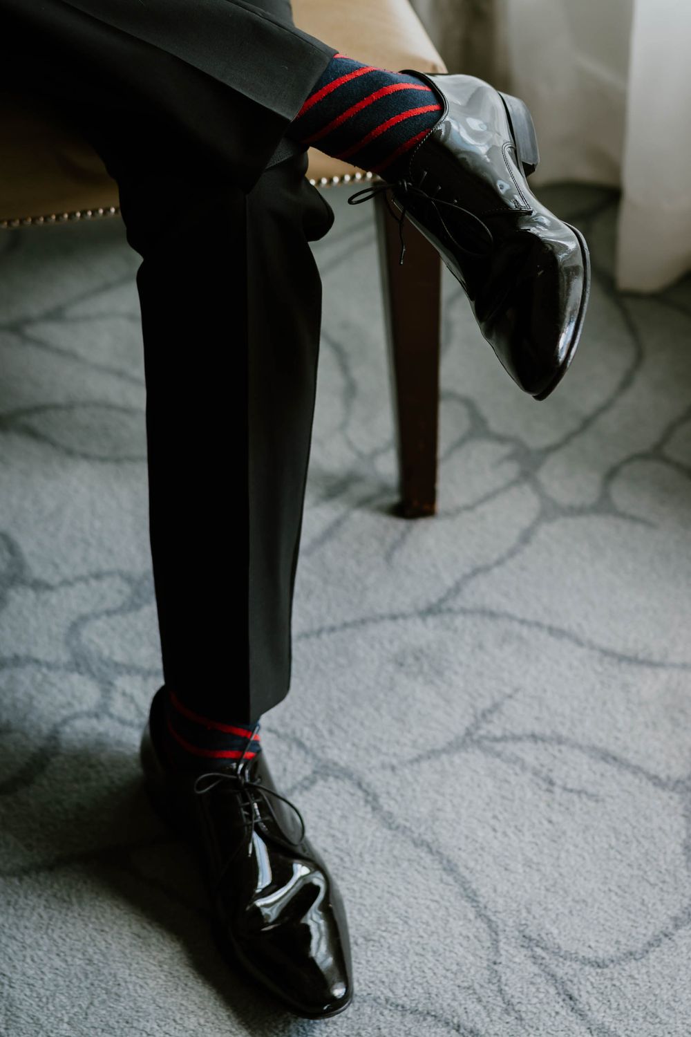 Man in black tuxedo pants wearing red and black striped socks with shiny patent leather dress shoes seated on chair.