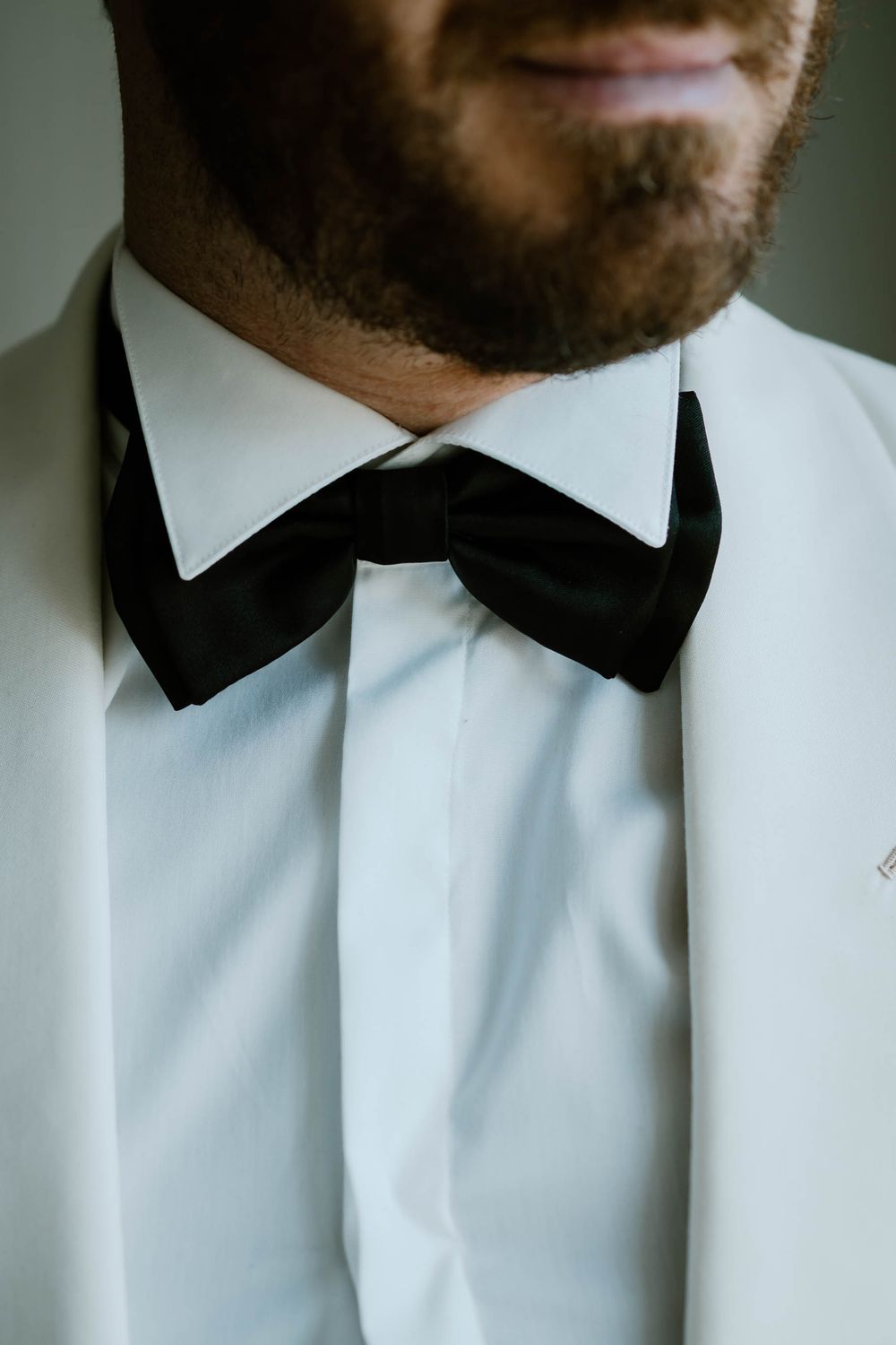 Groom in white tuxedo jacket with black bow tie and beard, close-up formal wedding attire detail photo.