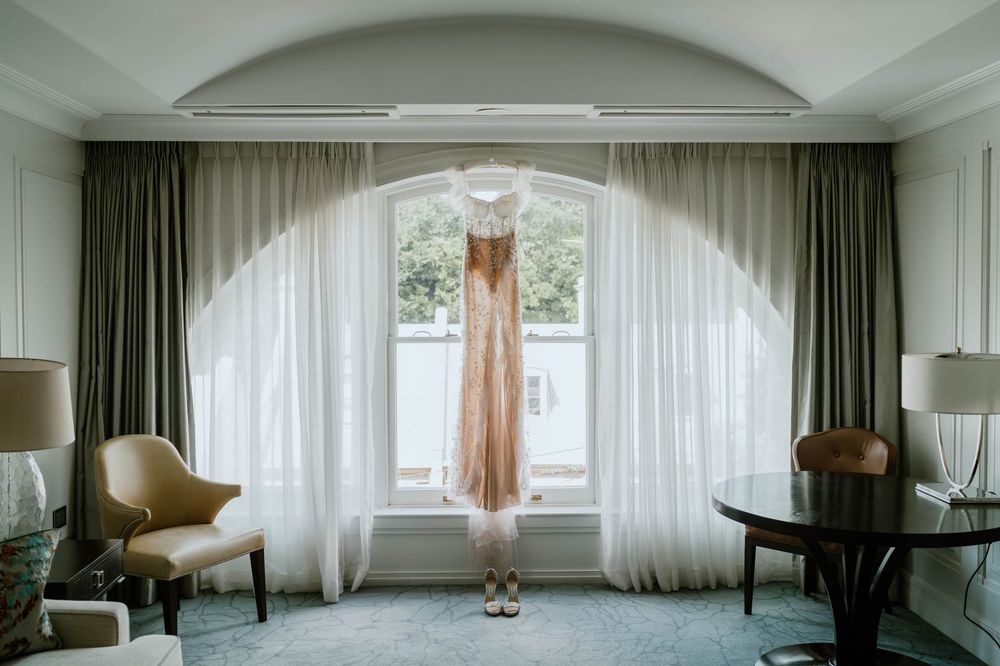A elegant rose gold wedding dress hanging in a luxurious hotel suite window with sheer white curtains.