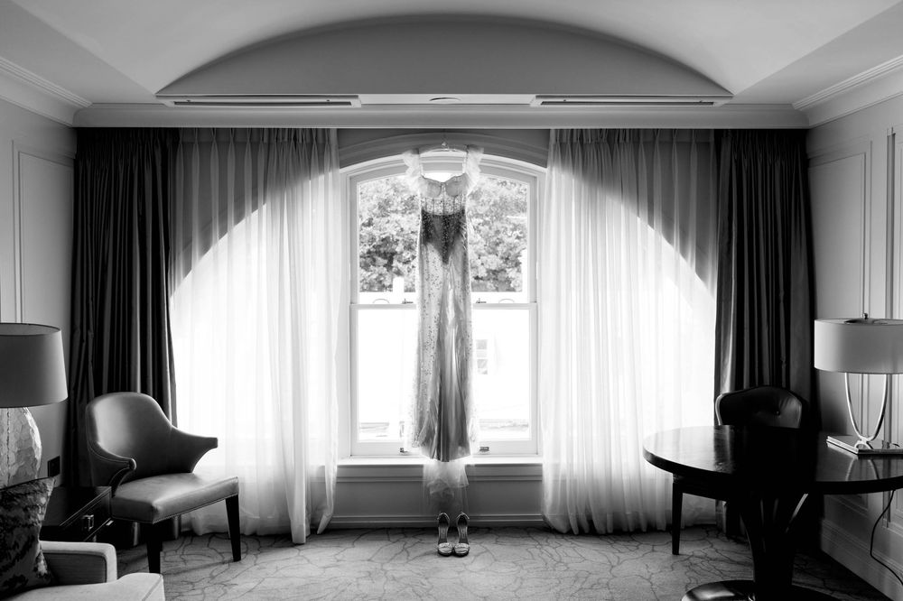 Black and white photo of an elegant wedding dress hanging in a hotel suite window with draped curtains.