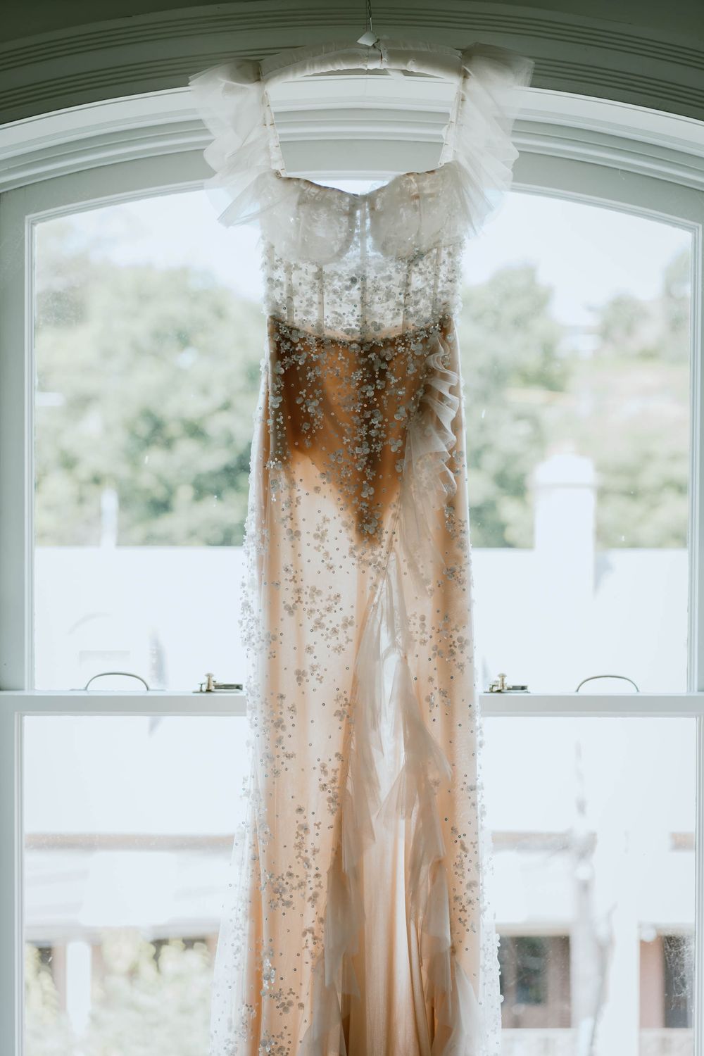 Elegant beaded champagne wedding gown hanging in arched window, featuring intricate crystal embellishments and lace details.