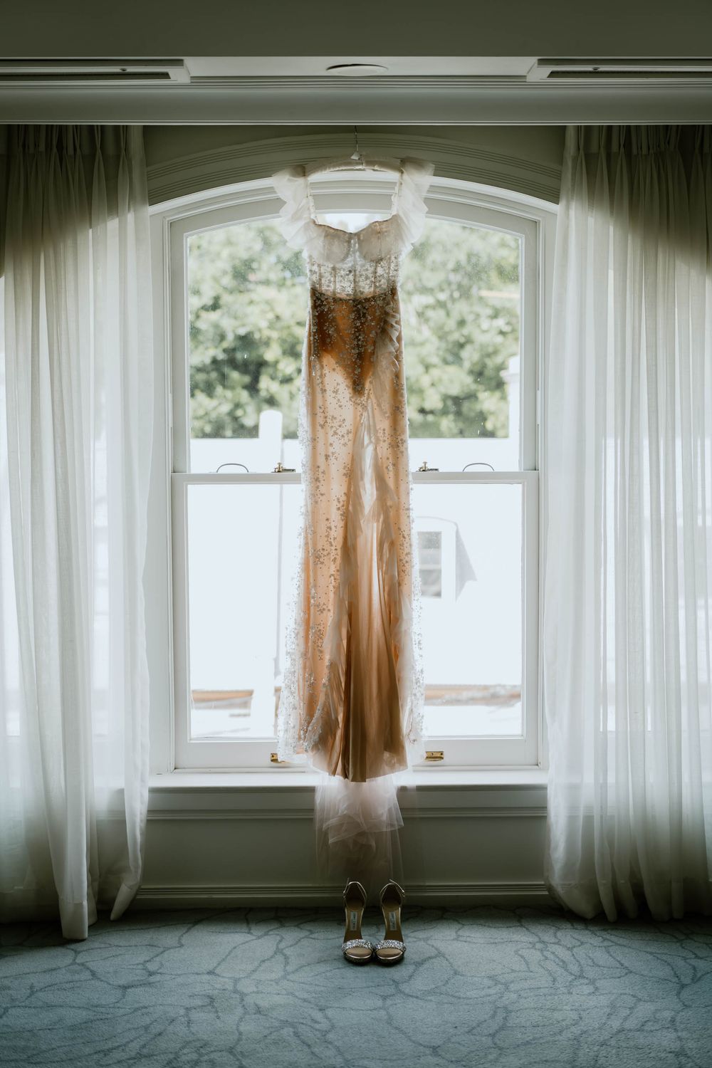Elegant gold beaded wedding dress hanging in arched window with matching heels below, soft natural light and sheer curtains.