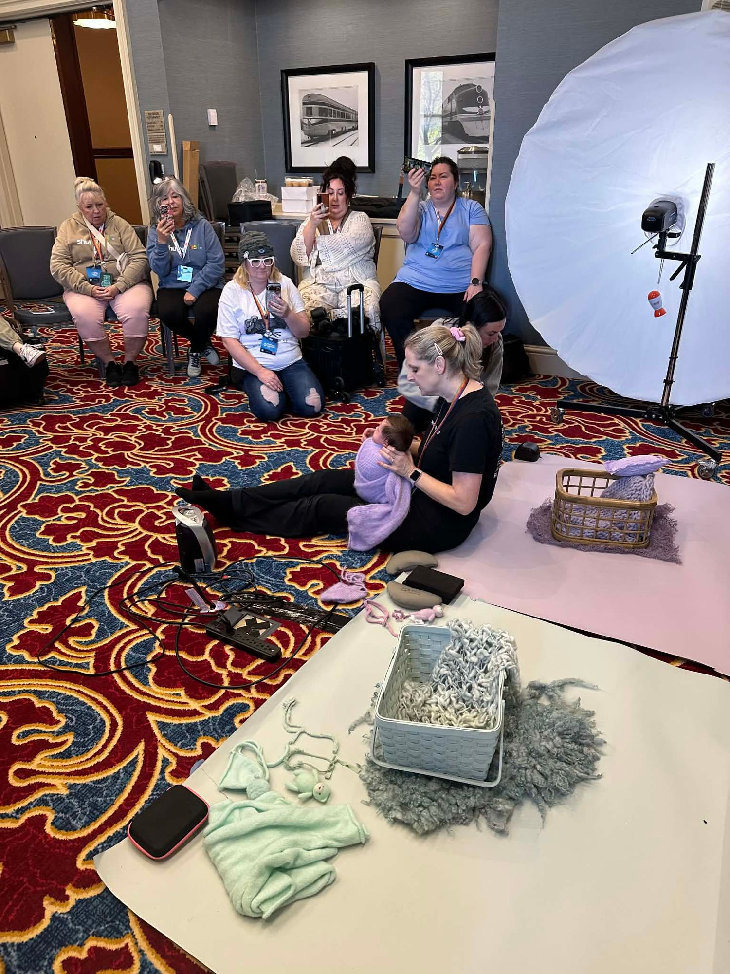 Photography workshop with attendees watching a demonstration using props, lighting umbrella, and newborn posing setup on patterned carpet.