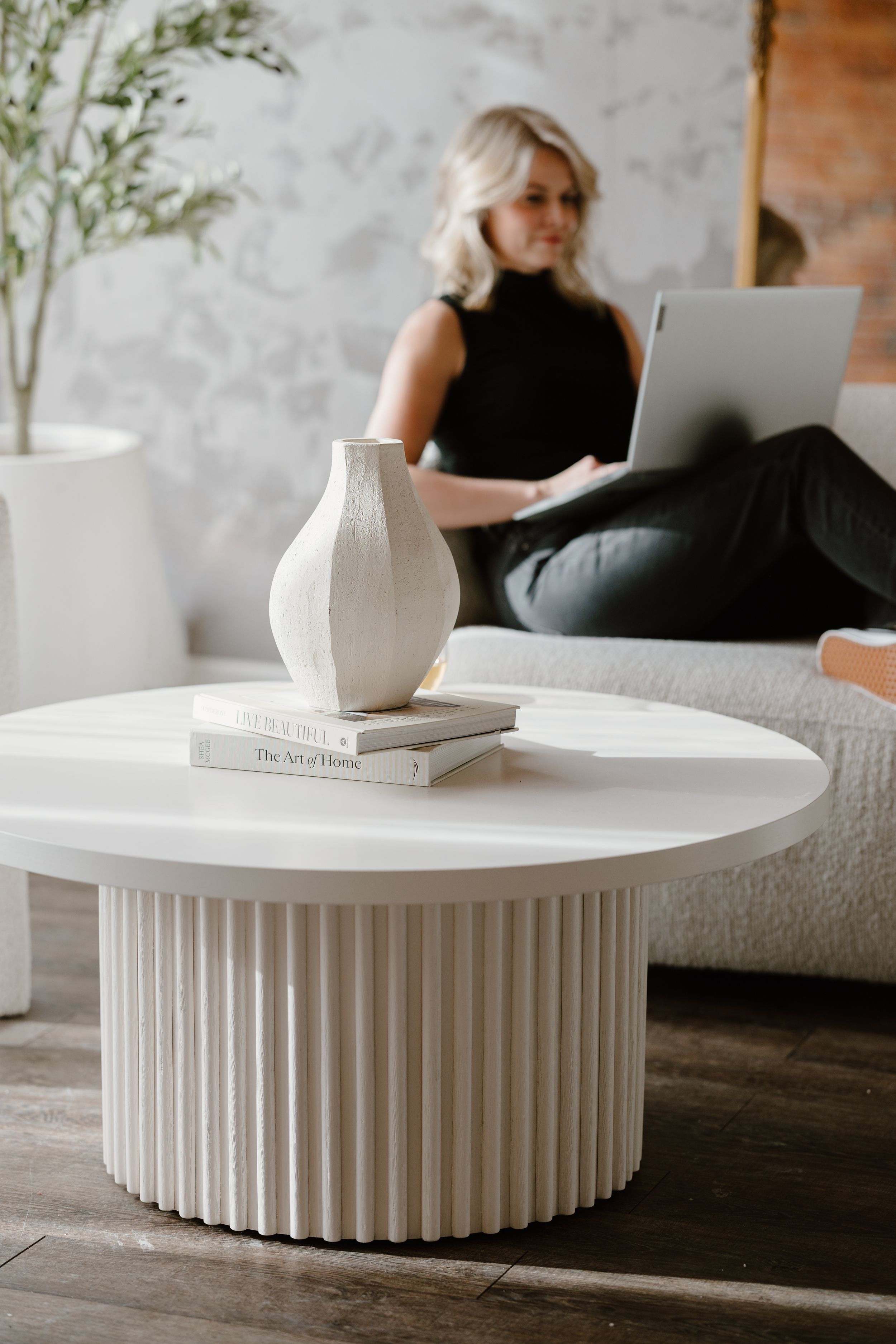 Woman working on laptop behind a white round fluted coffee table with a ceramic vase and books in a modern living room.
