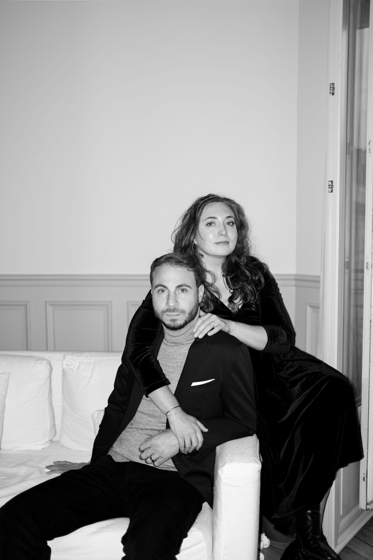 Black and white elegant photo of two people posed together on a white sofa in formal attire.