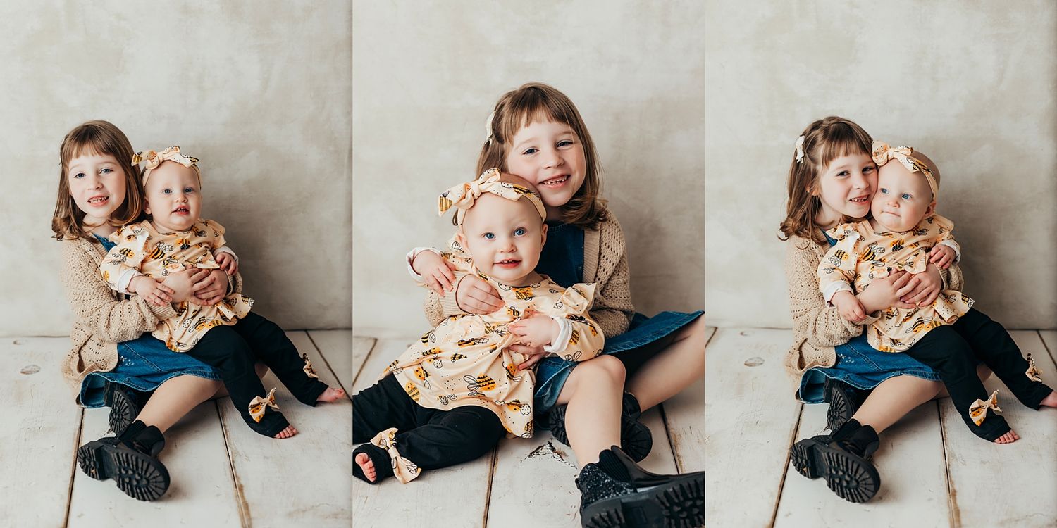 Two young sisters sitting together on the floor, older child hugging younger toddler, wearing matching floral outfits.