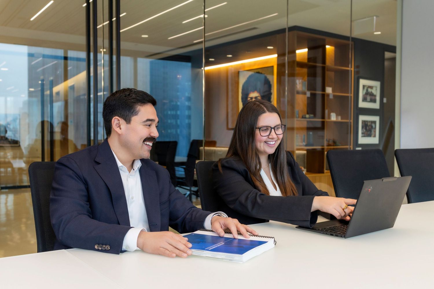 Two business professionals smiling and collaborating at a modern office table with a laptop and tablet.