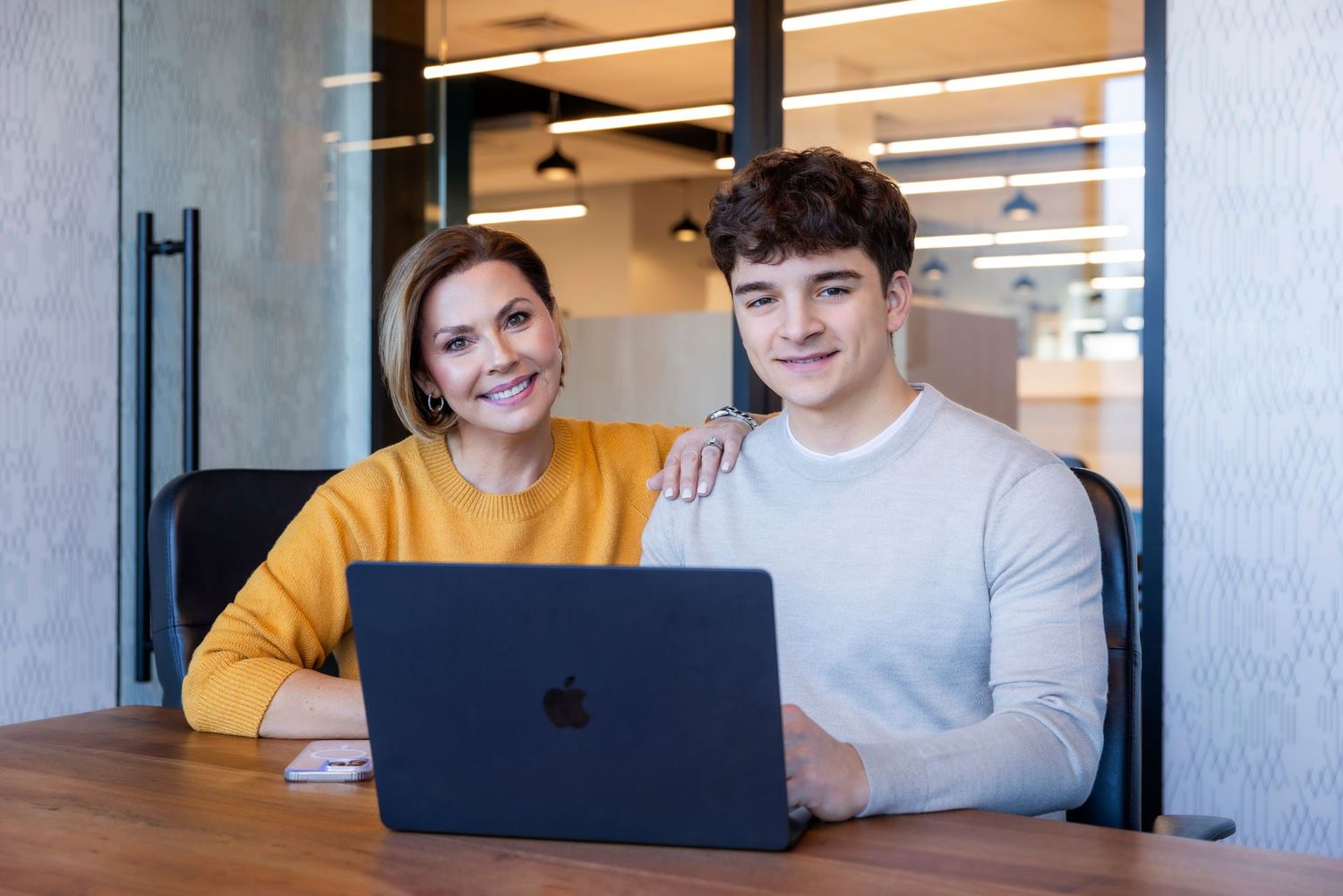Two colleagues smiling at camera while working together on a MacBook laptop at a wooden office desk.