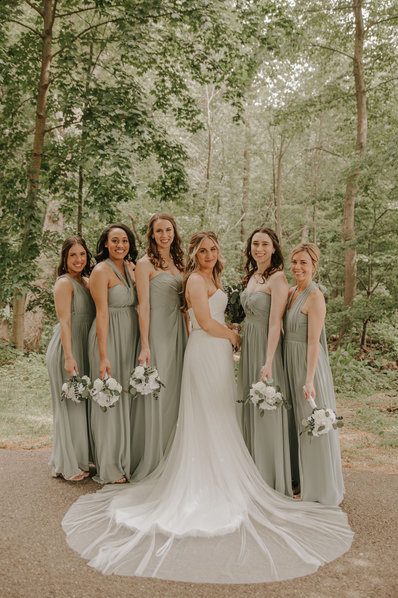 Bride in white gown with five bridesmaids in sage green dresses holding white bouquets in a wooded outdoor setting.