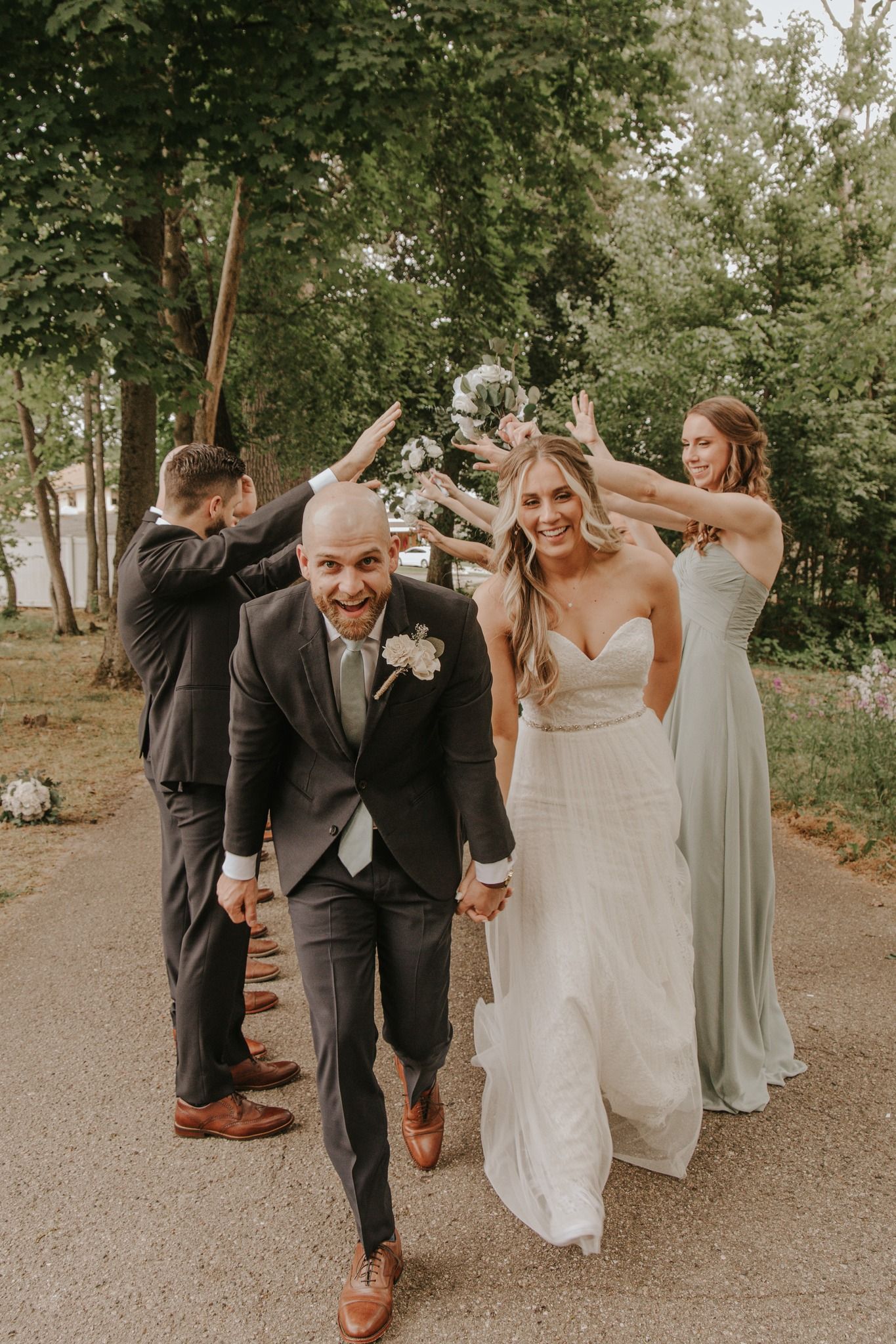 Joyful newlyweds walk hand-in-hand under raised arms of wedding party outdoors, bride in white gown, groom in dark suit.