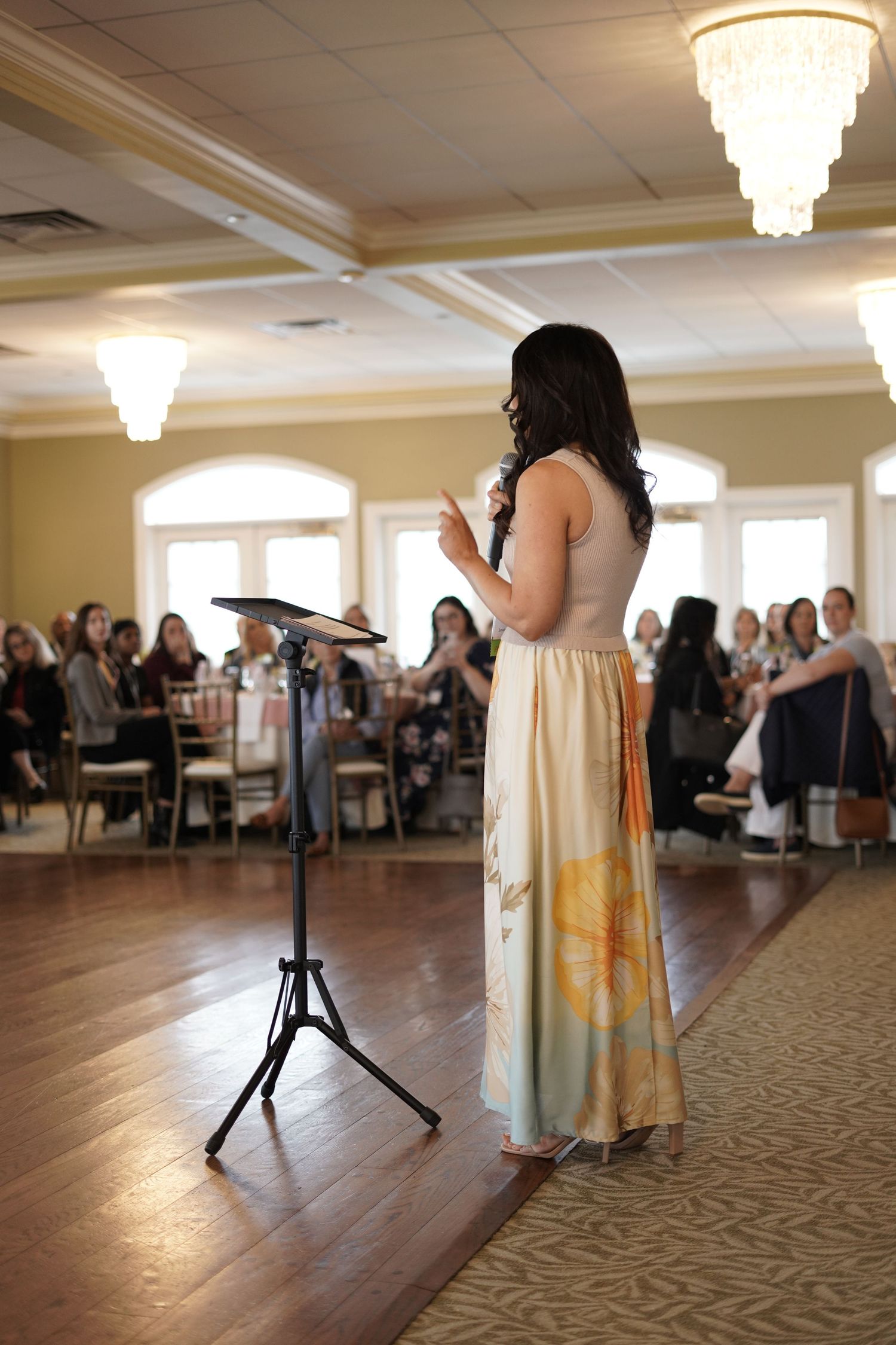Woman presenting at an elegant banquet hall event with seated audience and decorative displays.