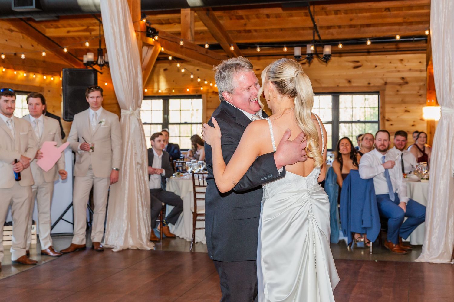 Bride and her father share a tender first dance at a rustic wedding reception with string lights and guests watching.