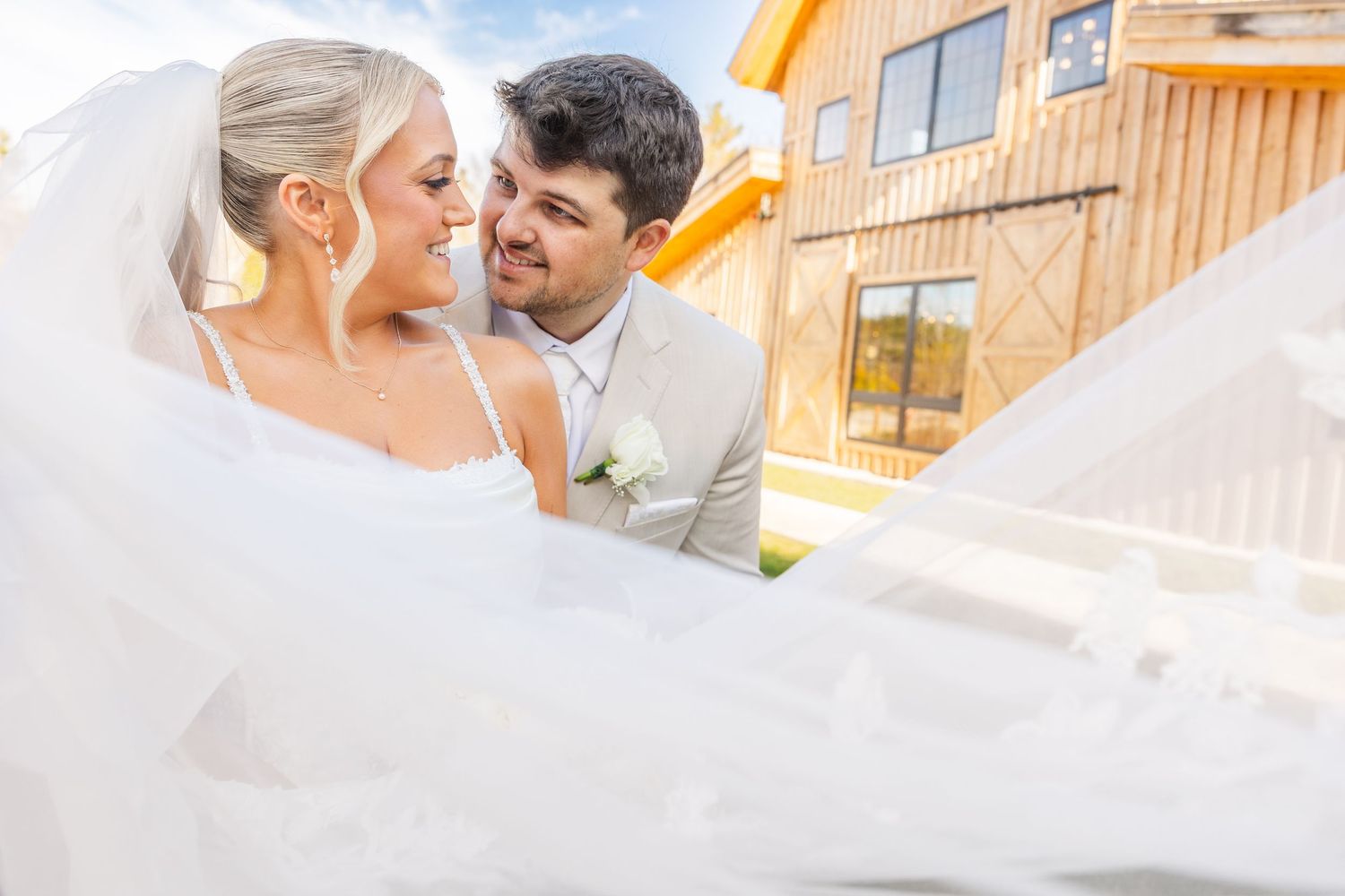Bride and groom share a romantic moment in front of a rustic barn venue, with wedding veil flowing in the breeze.