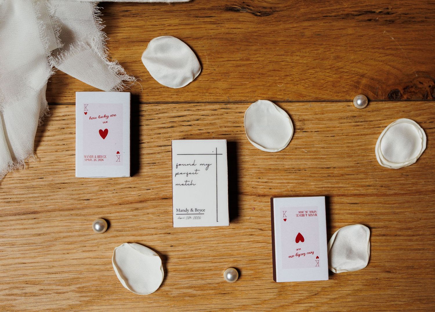 Wedding cards with red hearts and pearl accents arranged on a wooden surface with white flower petals.