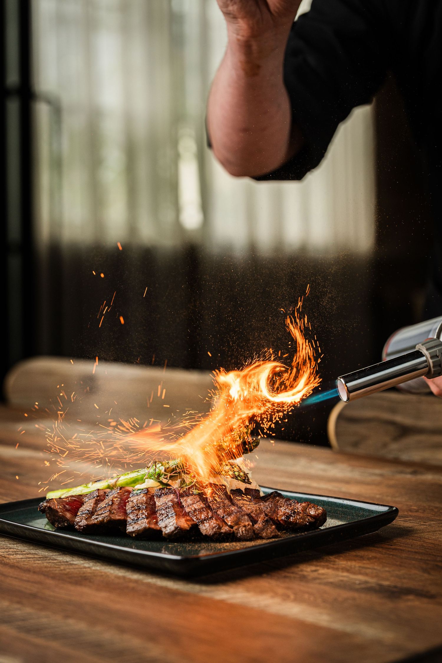 Chef using a blowtorch to flambé a sliced steak on a black plate, creating dramatic flames and sparks on a wooden table.