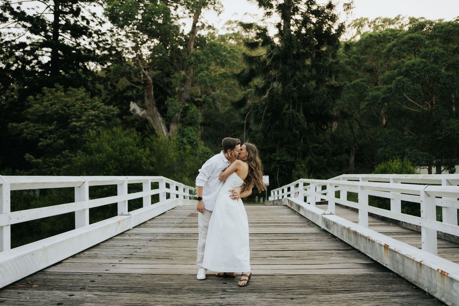 Couple shares a passionate kiss on a white wooden bridge surrounded by lush green trees.