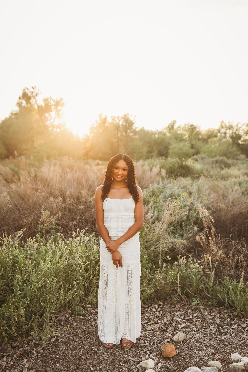 Young woman in white maxi dress standing in golden hour sunlight surrounded by lush greenery outdoors.
