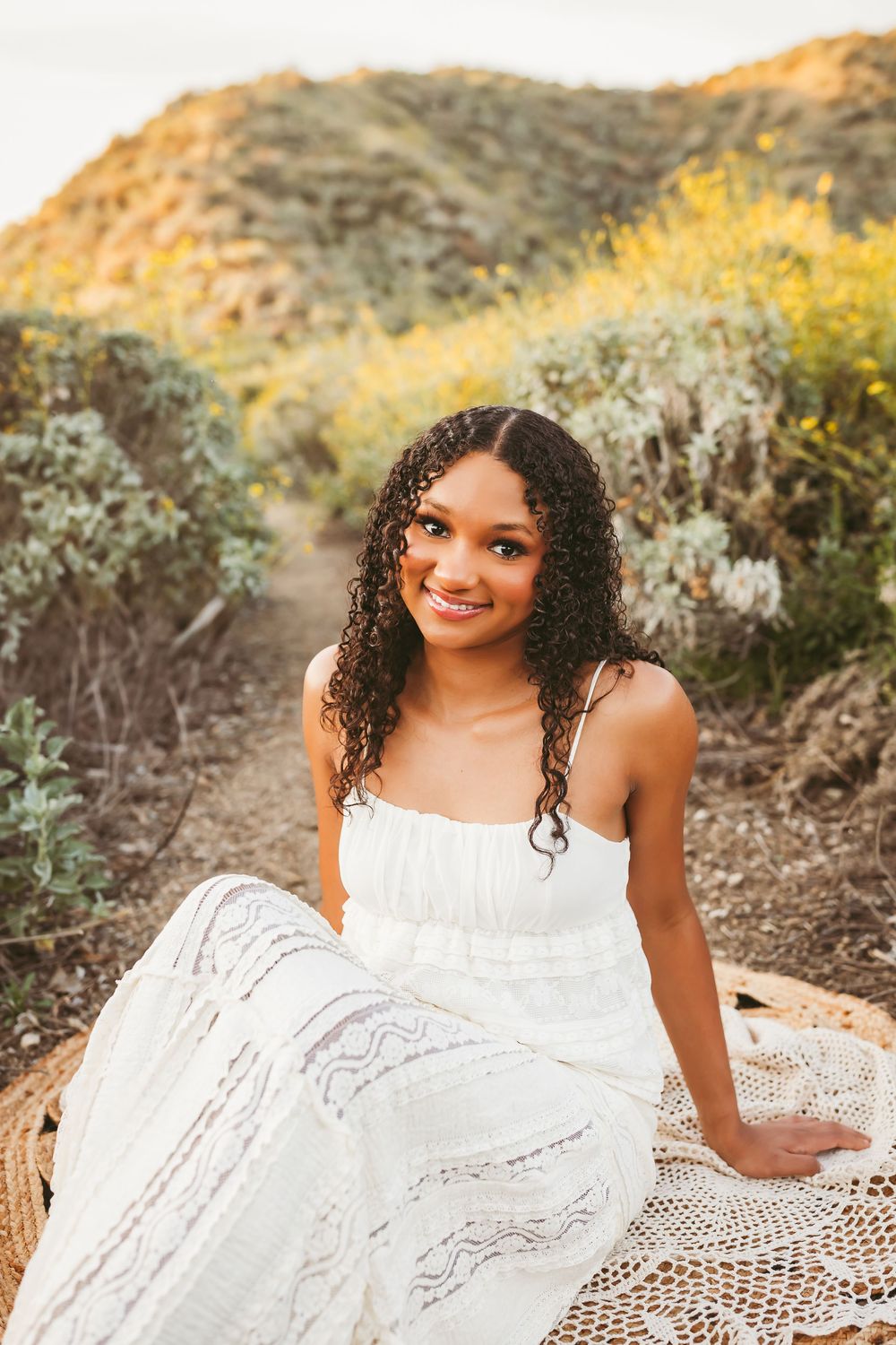 Young woman in white lace dress sitting outdoors on a trail surrounded by wildflowers and golden hills for her senior photos in redlands ca