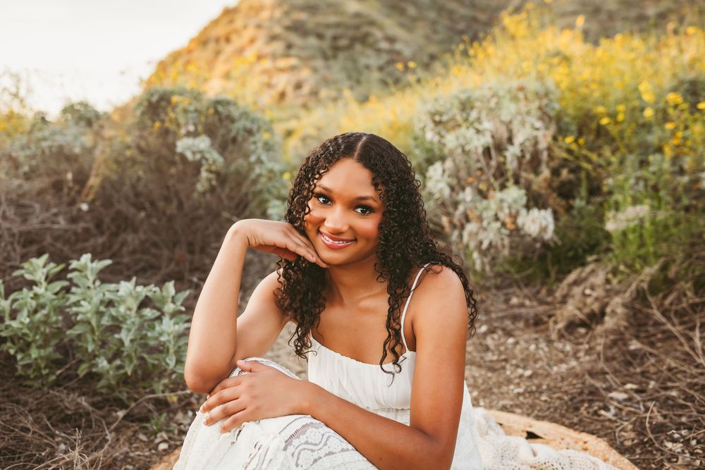 Young woman in white dress sitting outdoors among wildflowers with heidi grace photography for senior photos