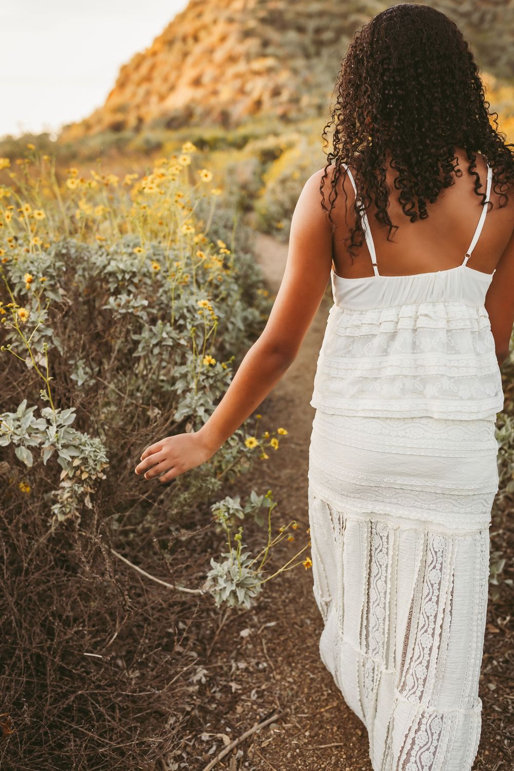 Woman in white lace dress walking through wildflower trail, touching yellow flowers with rocky hillside backdrop.