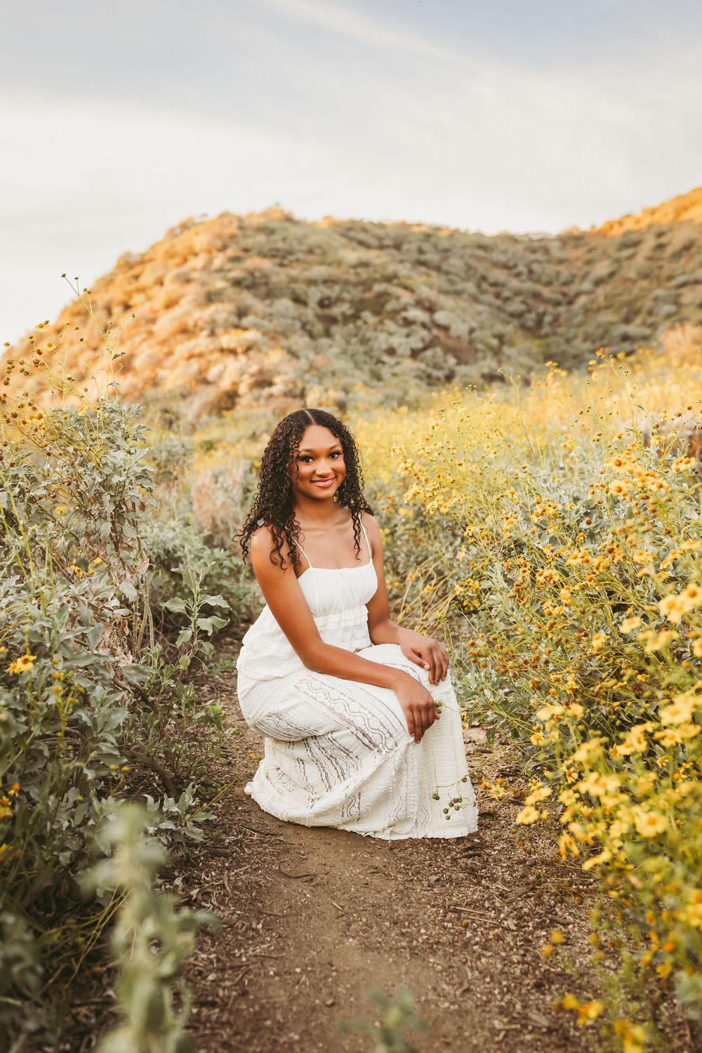 Young woman in white dress sitting on a dirt path surrounded by yellow wildflowers with a golden hillside backdrop.