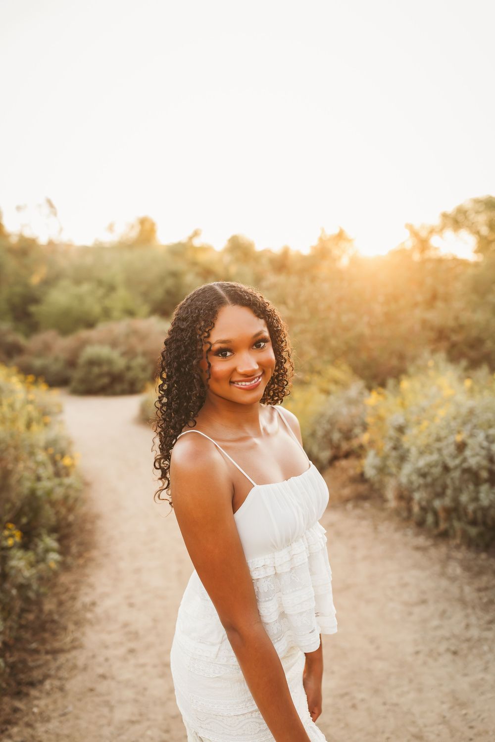 Young Black woman in white dress smiling on a sunlit garden path at golden hour.