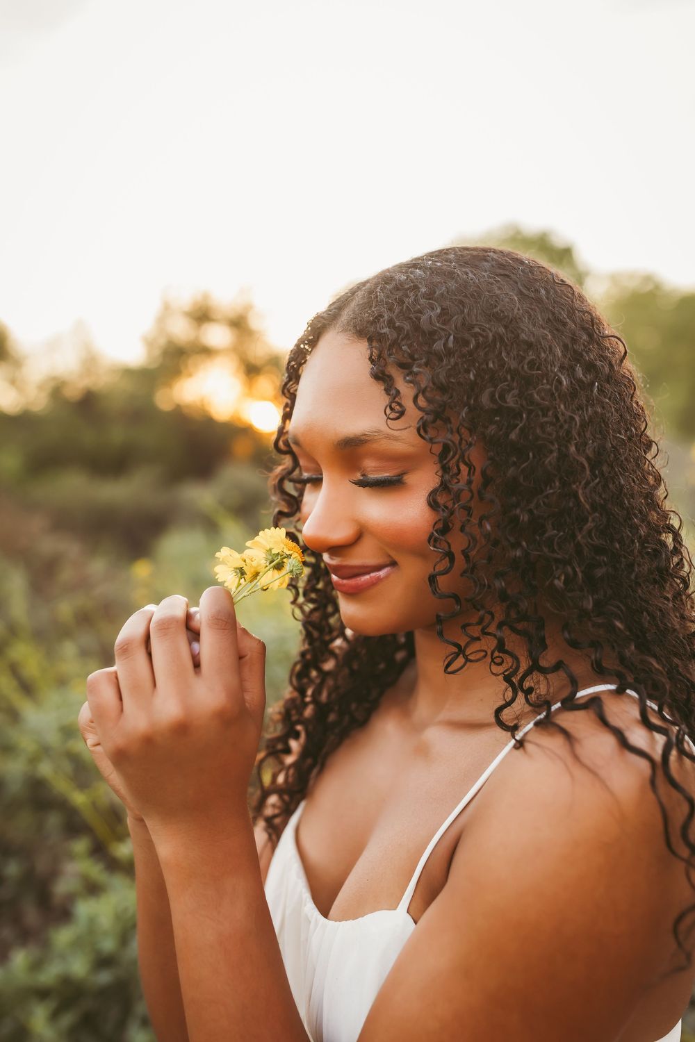 Young Black woman with curly hair smelling yellow wildflowers outdoors at golden hour in a white dress.