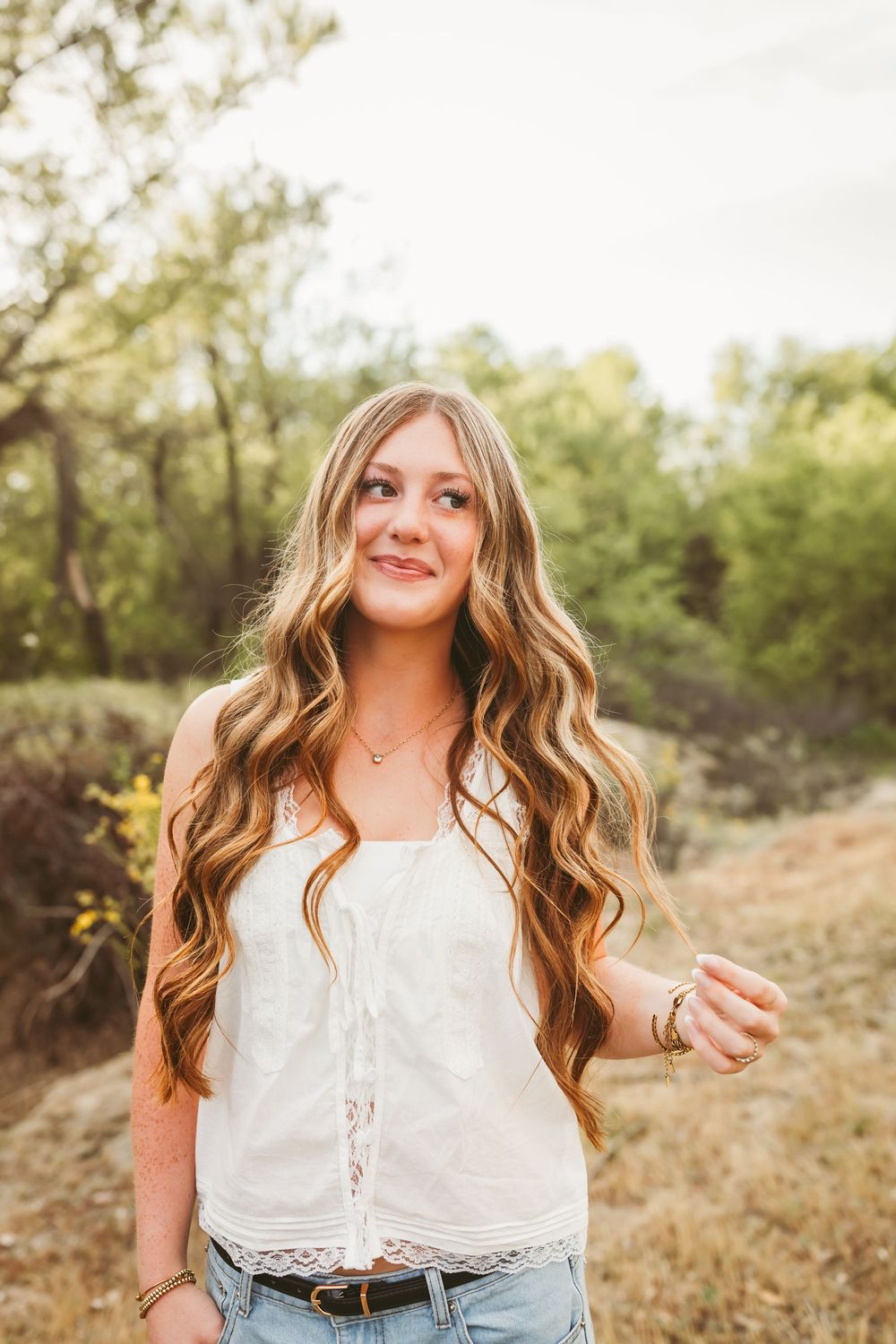Young woman with long wavy blonde hair wearing white lace top and jeans, smiling outdoors in natural setting.